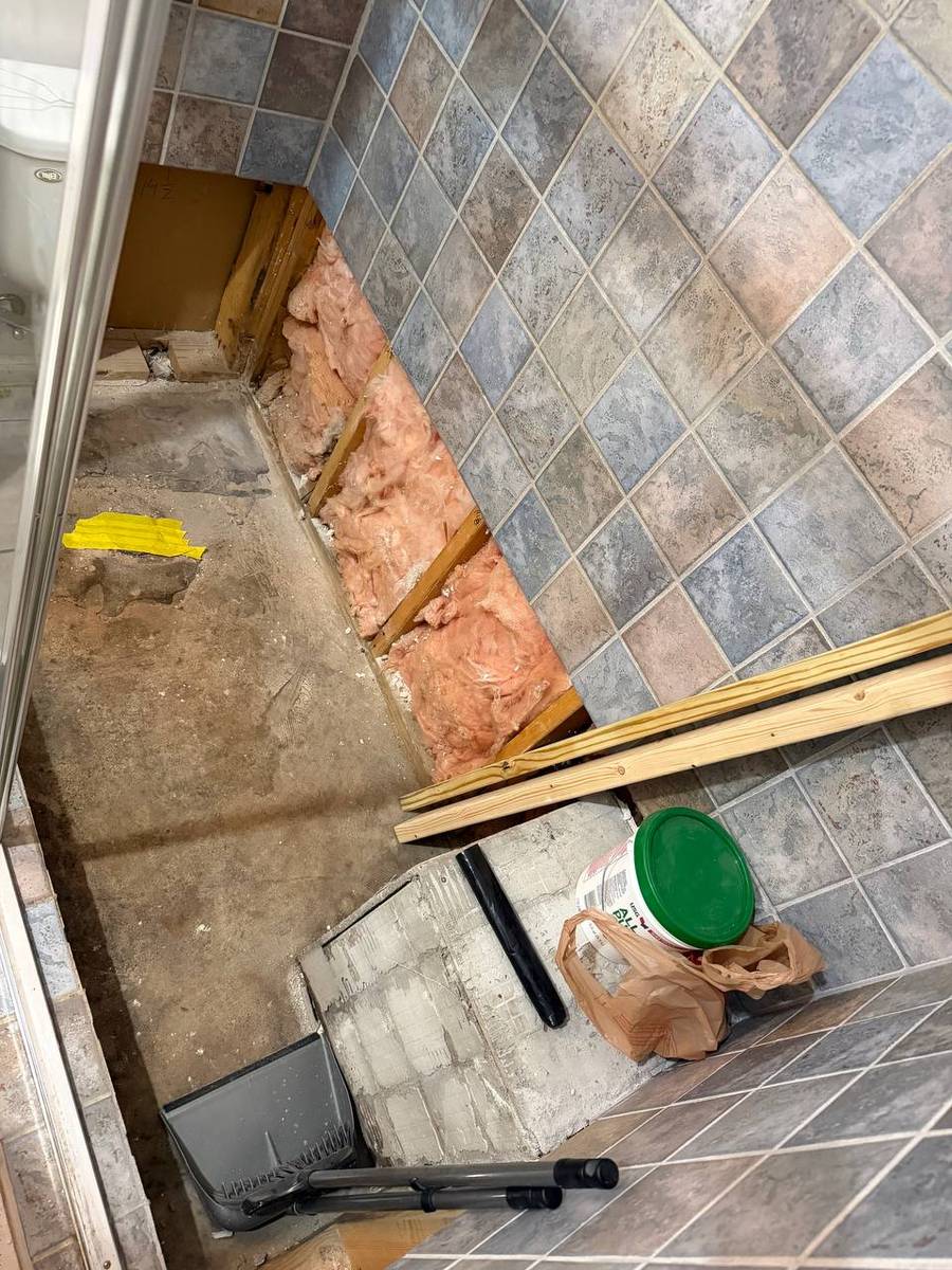 Demolished shower floor section down to subfloor and wall insulation, exposing the uneven concrete substrate causing standing water
